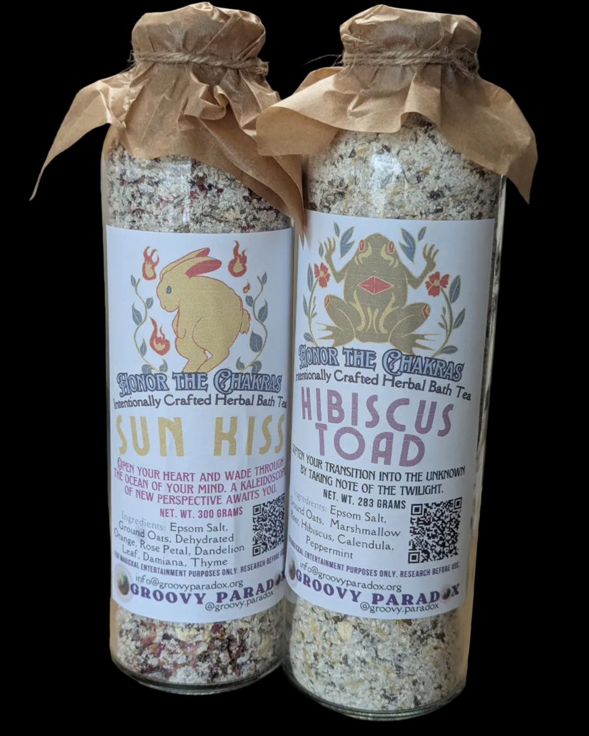Two tall glass jars filled with herbal bath salts, tied with brown paper wraps, and standing on a tabletop. The labels read ' 'Sun Kiss' and 'Hibiscus Toad' variants.