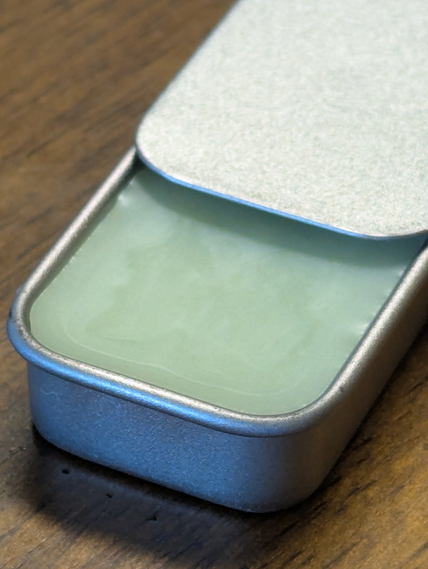 Close-up of a rectangular tin container with an open lid on a wooden surface. Inside the container is a green herbal mint lip balm.