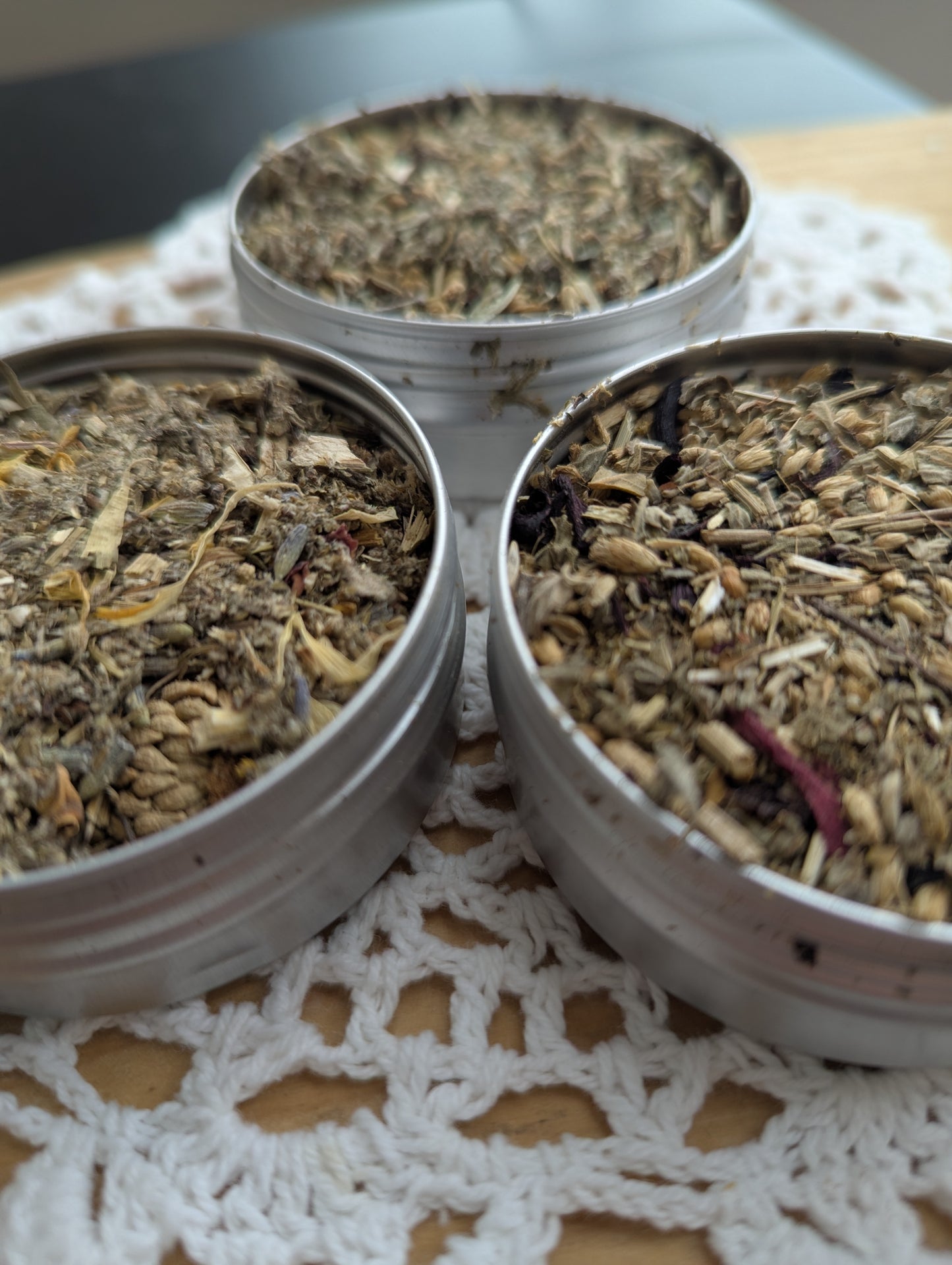 Three metal containers filled with different types of dried herbs on a lace tablecloth.