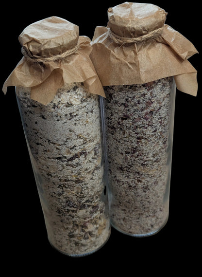 Two clear glass jars filled with a coarse, granular beige and brown mixture of Oat and Epsom Salt, sealed with brown corks and parchment paper tied around the necks, set on a flat surface.
