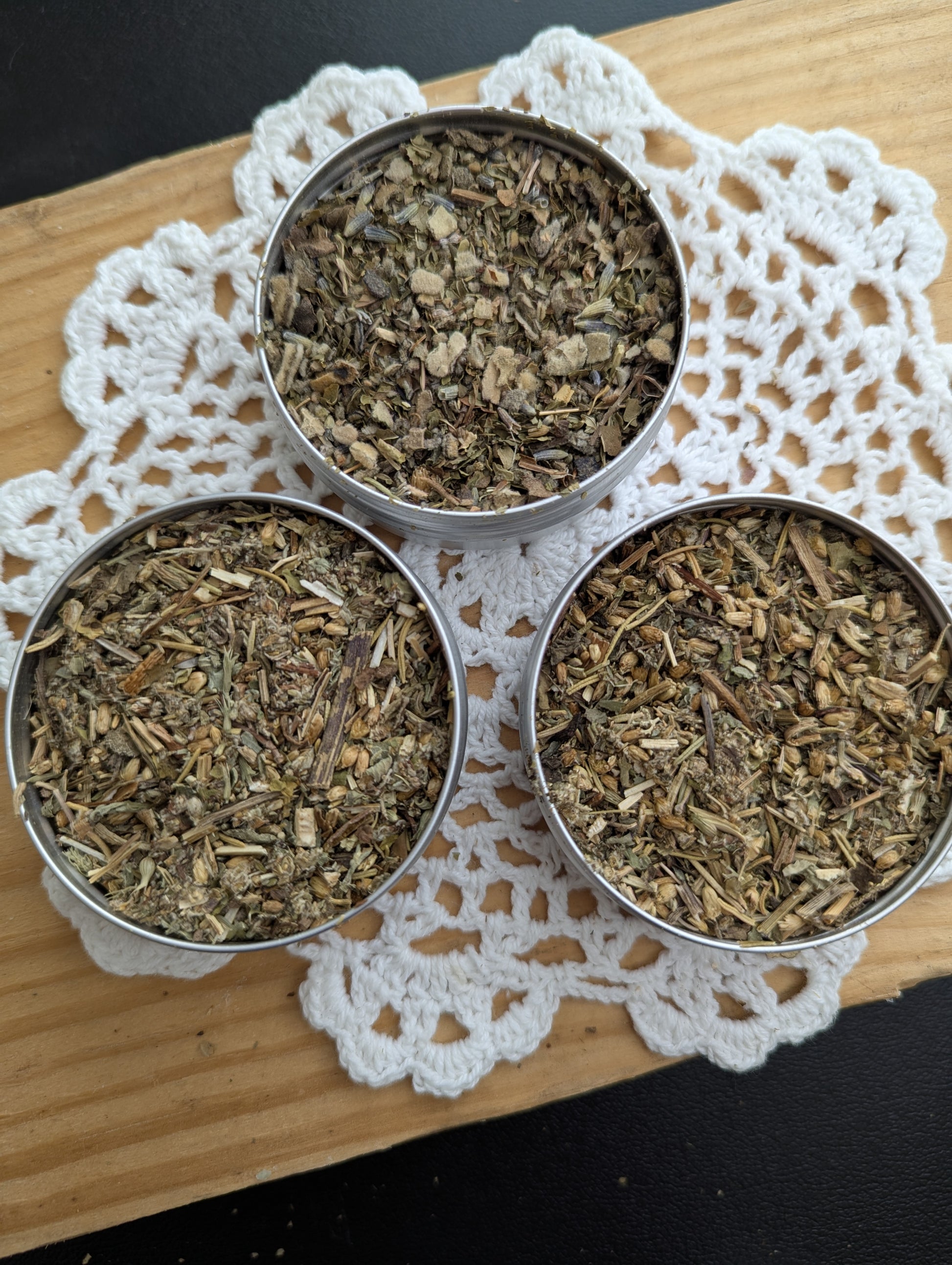 Three metal containers filled with dried herbs on a lace doily.