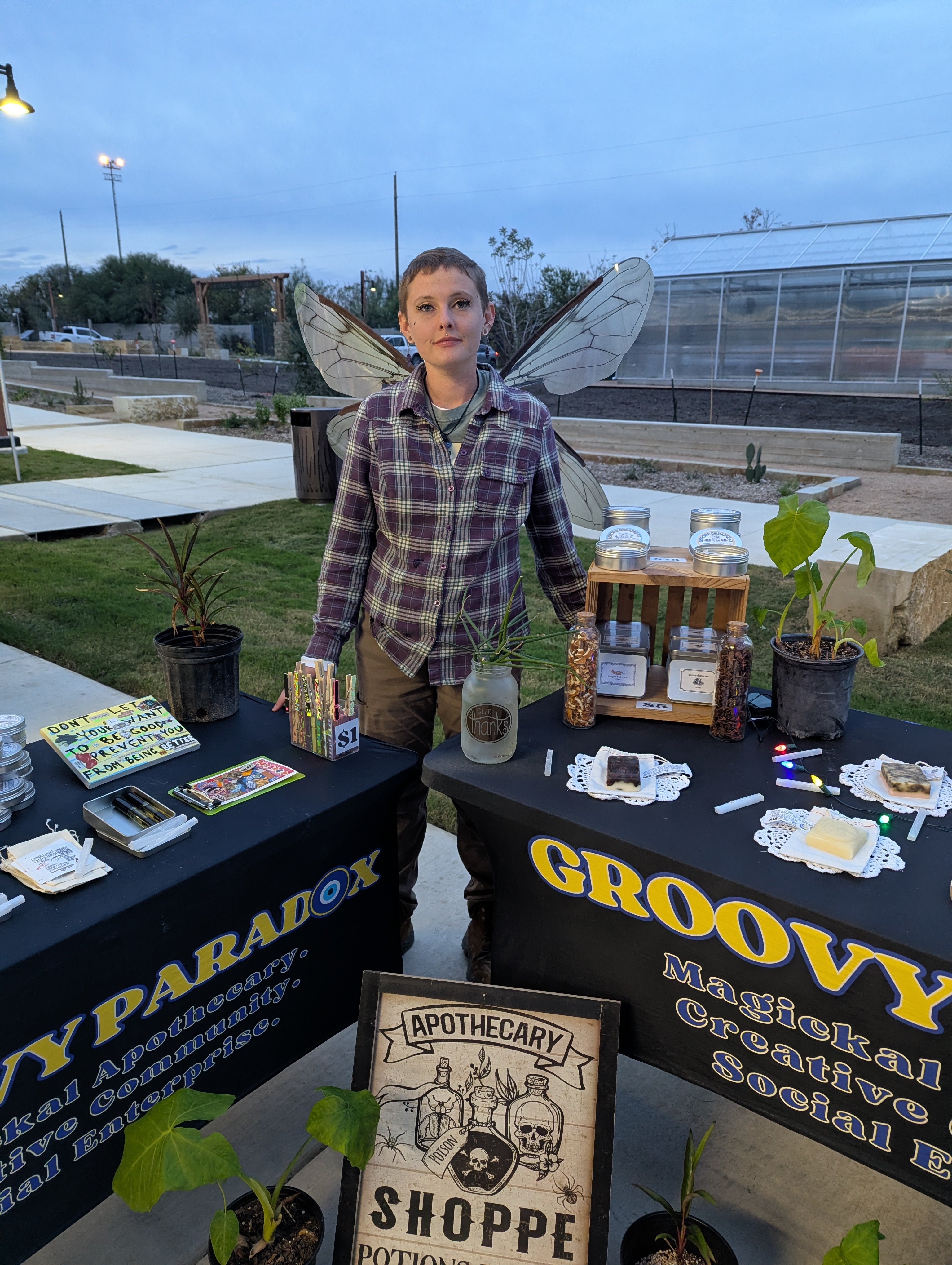 Person standing behind a table with various items at an outdoor event, including 'Groovy Apothecary Shoppe' branding.