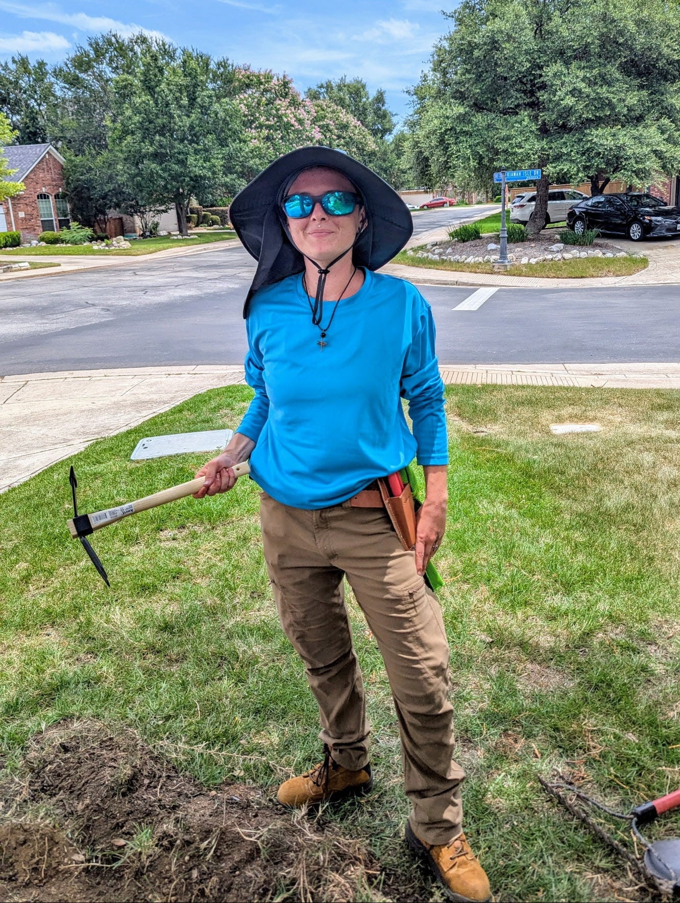 Person in blue shirt and hat holding gardening tools on grass with street and trees in background